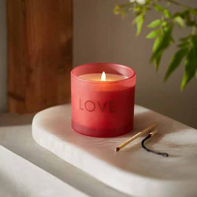 A red scented candle with the word 'Love' printed on it, placed on a white surface with a wooden background.